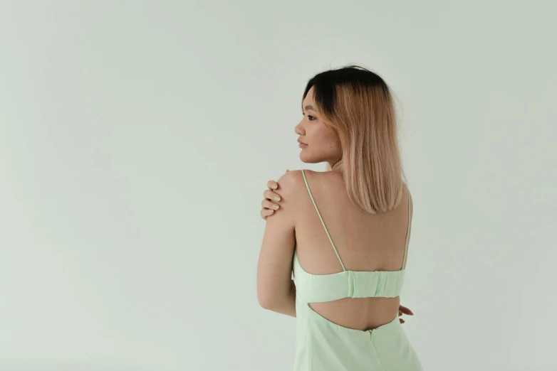 a woman in a green dress standing in front of a white wall, trending on pexels, aestheticism, posing in bed, young asian woman, bare back, pastel clothing