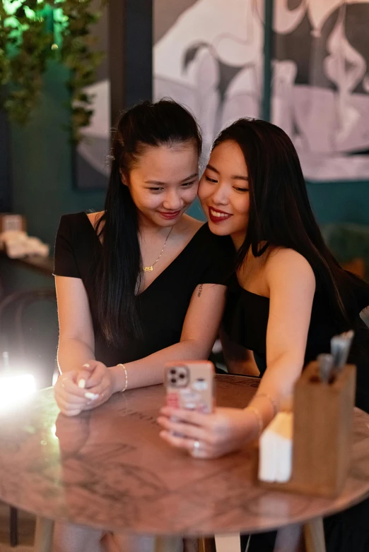 two women sitting at a table looking at a cell phone, a picture, inspired by Wang Duo, trending on pexels, beautiful asian girl, lesbian embrace, flashing lights, looking towards camera
