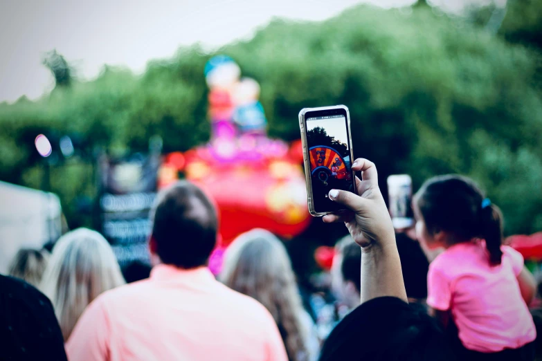 a person holding up a cell phone in front of a crowd, by Julia Pishtar, pexels contest winner, magic parade float, dreamworld, over the shoulder view, family friendly