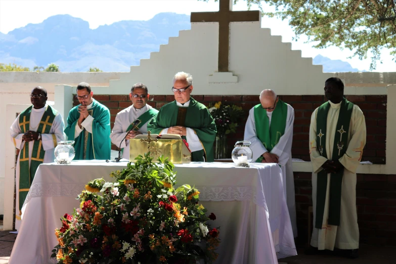 a group of men standing around a table in front of a cross, green robes, funeral, rosen zulu, giving a speech