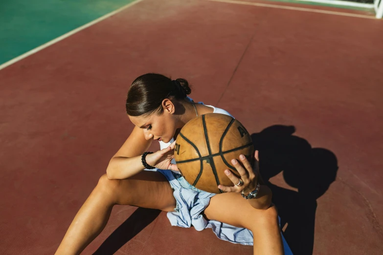 a woman sitting on the ground holding a basketball, tan skin, sunbathed skin, sport shorts, air shot