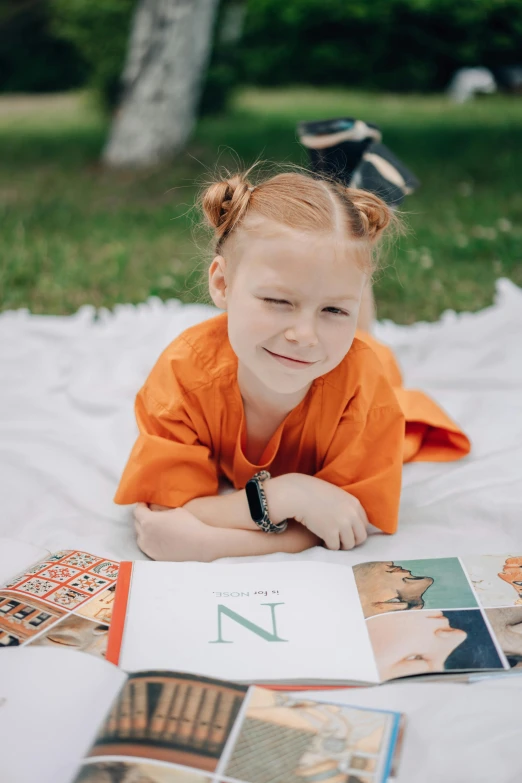 a little girl laying on a blanket reading a book, an album cover, unsplash, wearing an orange t shirt, yearbook photo, outdoor photo, uppercase letter