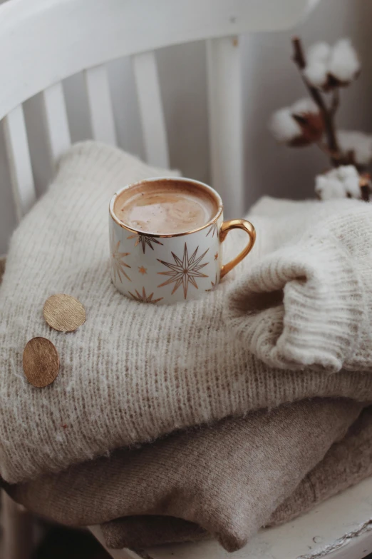 a cup of coffee sitting on top of a pile of sweaters, by Lucia Peka, trending on pexels, romanticism, coffee and stars background, subtle gold accents, botanicals, pregnancy