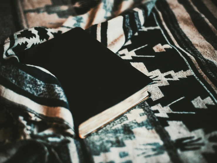 a book sitting on top of a blanket on top of a bed, by Carey Morris, pexels contest winner, hurufiyya, black cloth, an unknown ethnographic object, retro stylised, panel of black
