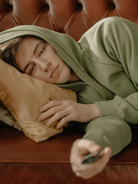 a person laying on a couch with a remote control, a colorized photo, inspired by Elsa Bleda, trending on pexels, hyperrealism, gray hoodie, sad grumpy face, wearing green jacket, joe keery