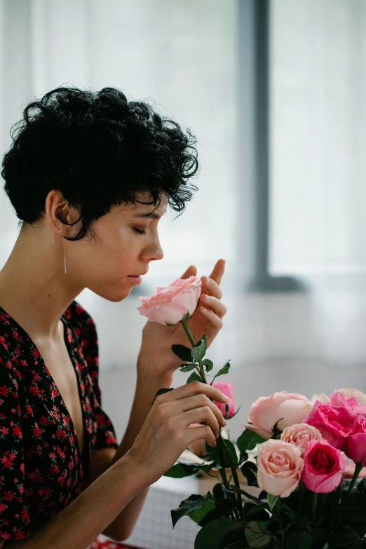 a woman sitting at a table with a vase of flowers, curly pixie cut hair, holding a rose in a hand, woman's face looking off camera, 2019 trending photo