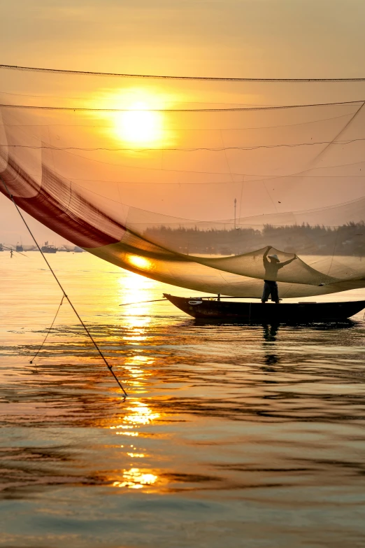 a man that is standing in the water with a net, flickr, vietnam, sun setting, silk flowing in wind, boat