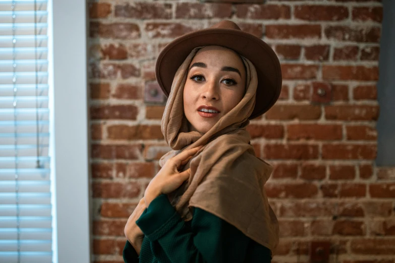 a woman standing in front of a brick wall, a portrait, inspired by Altoon Sultan, trending on pexels, hurufiyya, caracter with brown hat, asian human, scarf, emerald