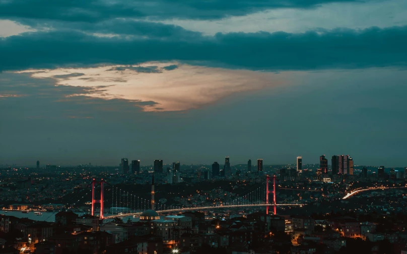 a view of a city at night from the top of a hill, inspired by Elsa Bleda, pexels contest winner, hurufiyya, turkish and russian, background image, on a cloudy day, tall bridge with city on top