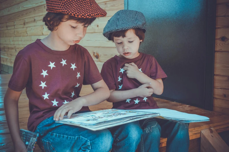 two young boys sitting on a bench reading a book, by Lucia Peka, pexels contest winner, graphic tees, black beret with a red star, jeans and t shirt, maroon hat