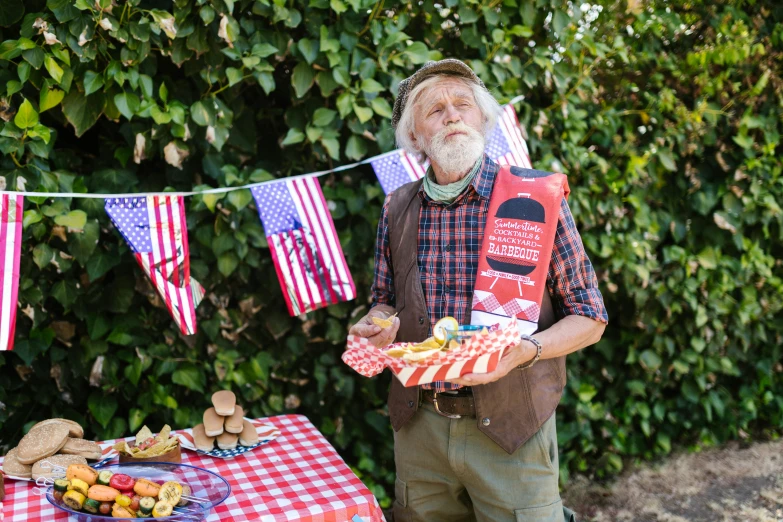 a man standing next to a table with food on it, inspired by Robert Ballagh, pexels, folk art, 4th of july, holding a red banner, emma bridgewater and paperchase, overalls and a white beard