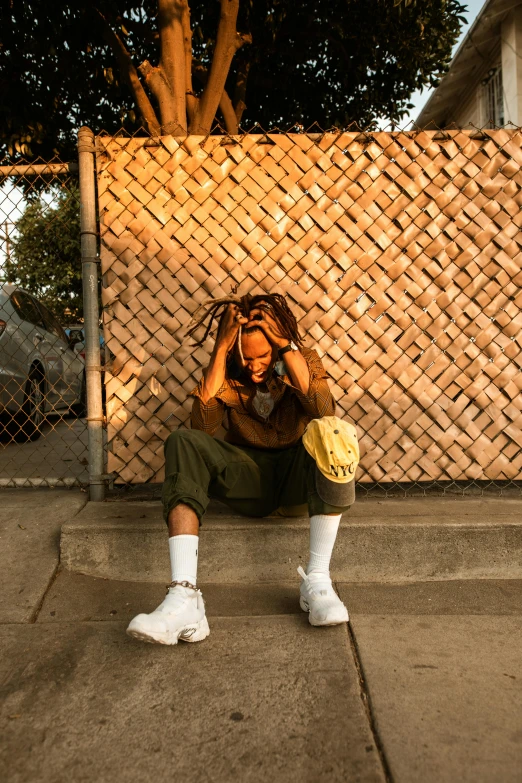 a man sitting on the sidewalk in front of a fence, box braids, hear no evil, golden glow, wearing a baggy