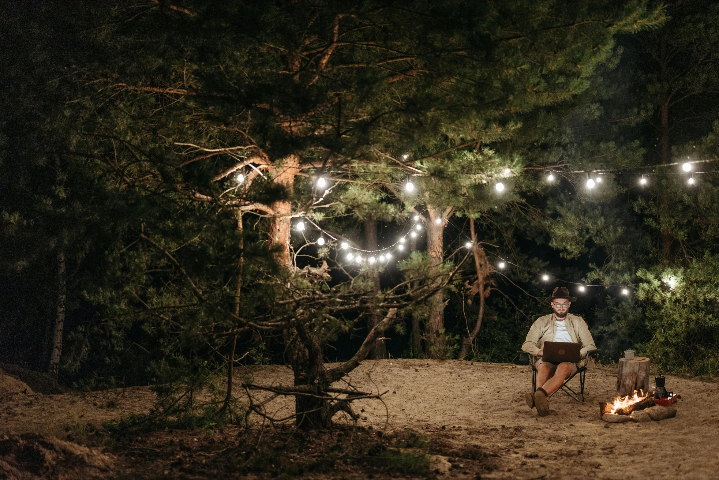 a woman sitting in a chair next to a fire, by Emma Andijewska, pexels contest winner, in a candy forest! at night, forest picnic, string lights, a handsome