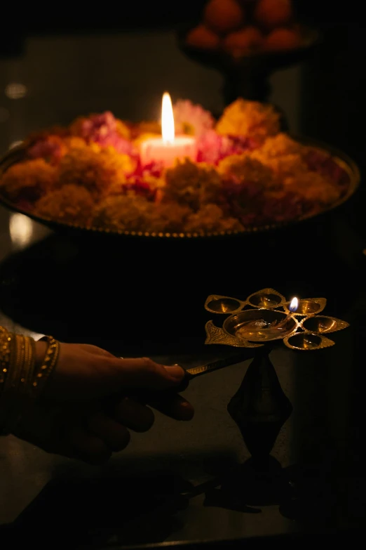 a person holding a lit candle in front of a plate of food, hindu ornaments, lamps and flowers, lit up in a dark room