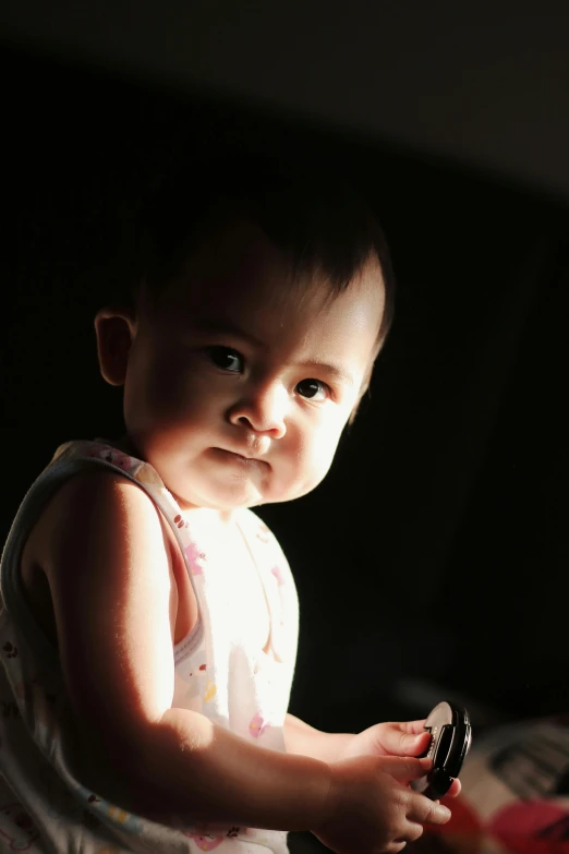 a baby sitting on top of a bed holding a remote, a portrait, by Basuki Abdullah, flickr, standing under a beam of light, looking serious, backlight glow, with a black background