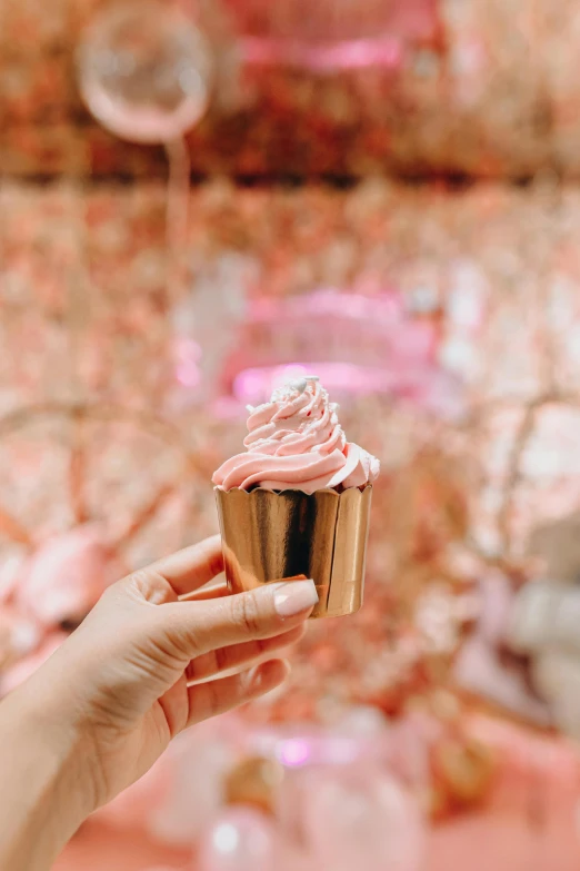 a person holding a cupcake with pink frosting, rose gold, complex background, instagram picture, promo image