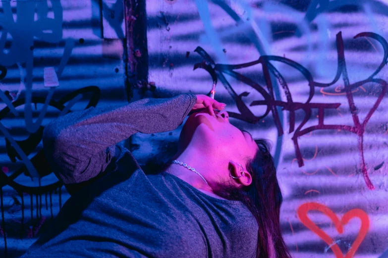 a woman smoking a cigarette in front of a graffiti covered wall, inspired by Elsa Bleda, pexels contest winner, neon light showing injuries, lying down, blue and purle lighting, strangled with rope