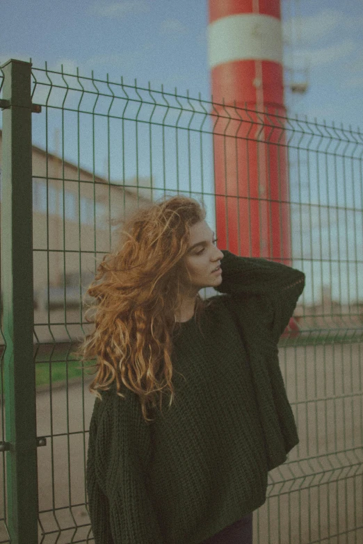 a woman leaning against a fence with a lighthouse in the background, inspired by Elsa Bleda, ginger wavy hair, woman in streetwear, desaturated, wearing a green sweater