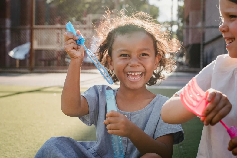 a couple of young girls sitting on top of a lush green field, pexels contest winner, process art, holding a blue lightsaber, she is smiling and excited, iridescent soapy bubbles, mixed race