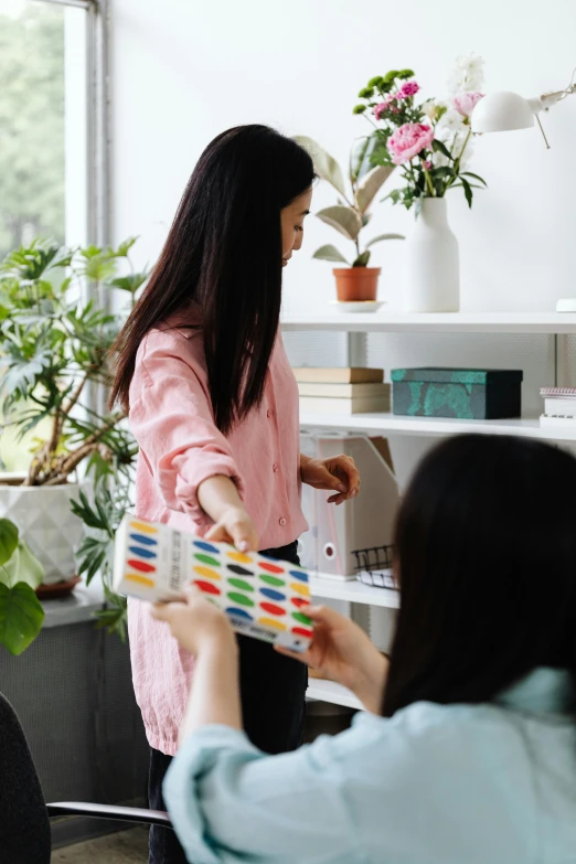 a woman that is standing in front of a desk, playing board games, collection product, next to a plant, coworkers