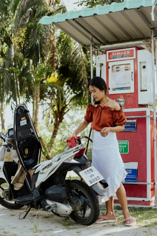 a woman standing next to a motorcycle at a gas station, pexels contest winner, white skirt and barechest, bali, square, gif