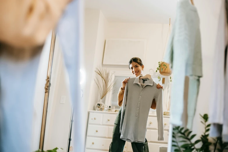 a woman standing in front of a mirror holding a shirt, by Nicolette Macnamara, pexels contest winner, happening, long shirt, a still of a happy, grey, thumbnail