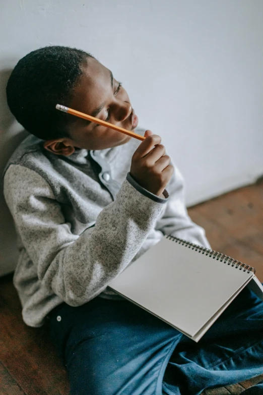 a young boy sitting on the floor with a notebook and pencil, trending on pexels, visual art, african american young woman, lost in thought, androgynous male, over his shoulder