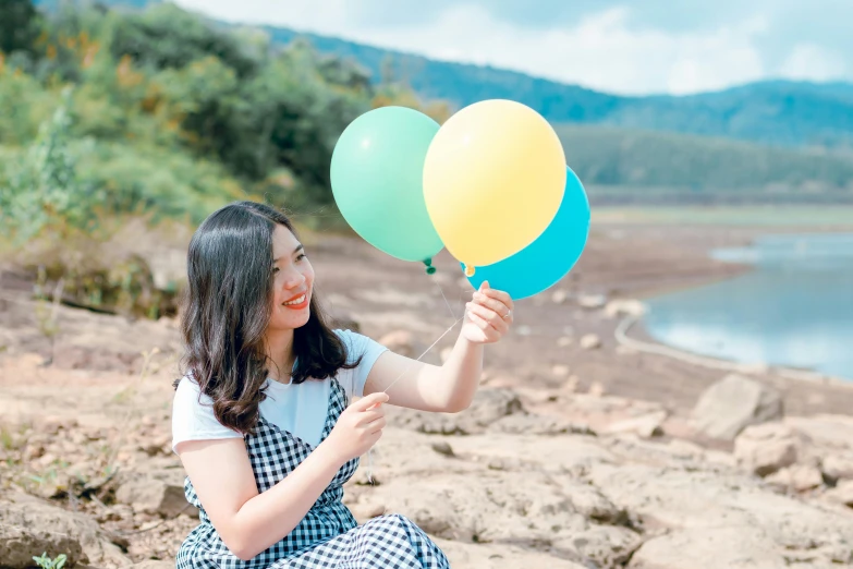 a woman sitting on a rock holding a bunch of balloons, inspired by Kim Jeong-hui, pexels contest winner, yellow and blue and cyan, happy friend, ulzzang, avatar image