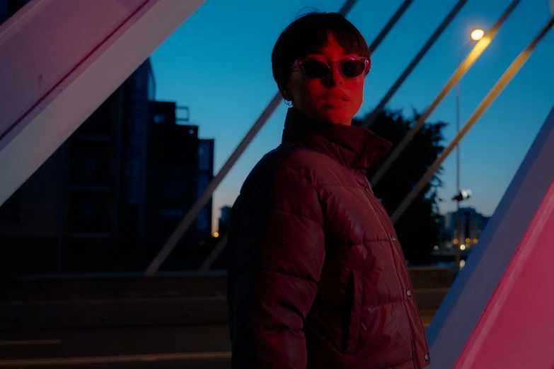 a woman standing in front of a bridge at night, an album cover, inspired by Zhu Da, pexels contest winner, model wears a puffer jacket, woman with rose tinted glasses, at future neon light rooftop, red and cinematic lighting