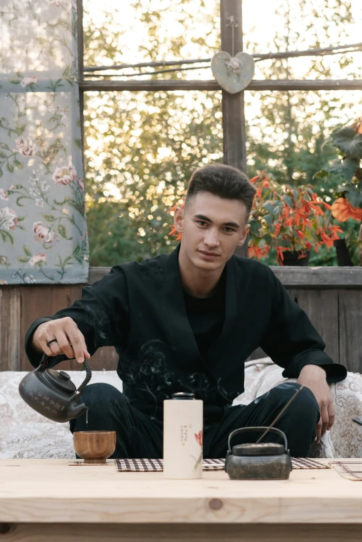 a man sitting on top of a wooden table, an album cover, by Alexander Runciman, trending on unsplash, mannerism, tea ceremony scene, cute young man, ukrainian, non binary model