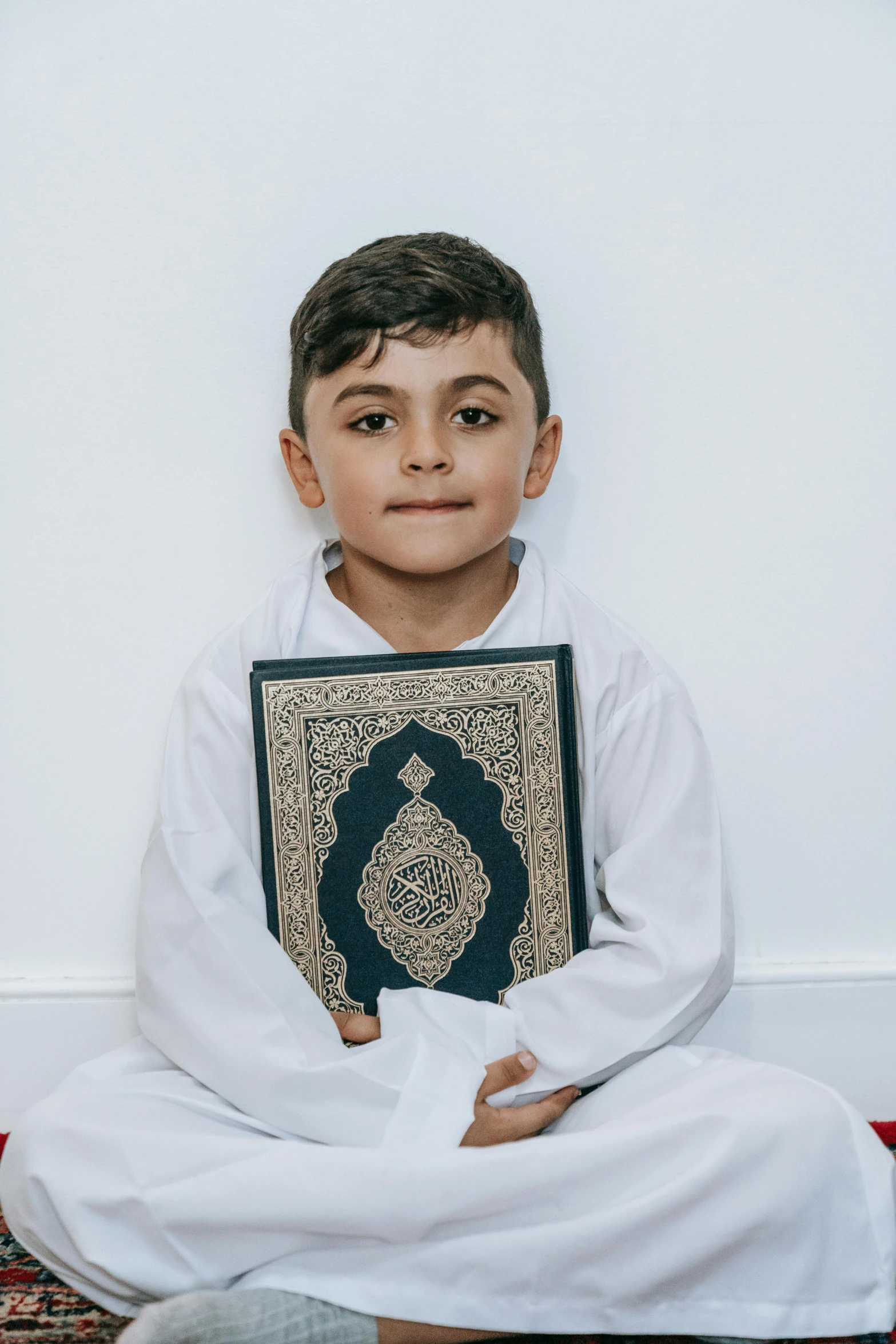 a young boy sitting on the floor holding a book, inspired by Sheikh Hamdullah, face centered, paisley, wearing white cloths, navy