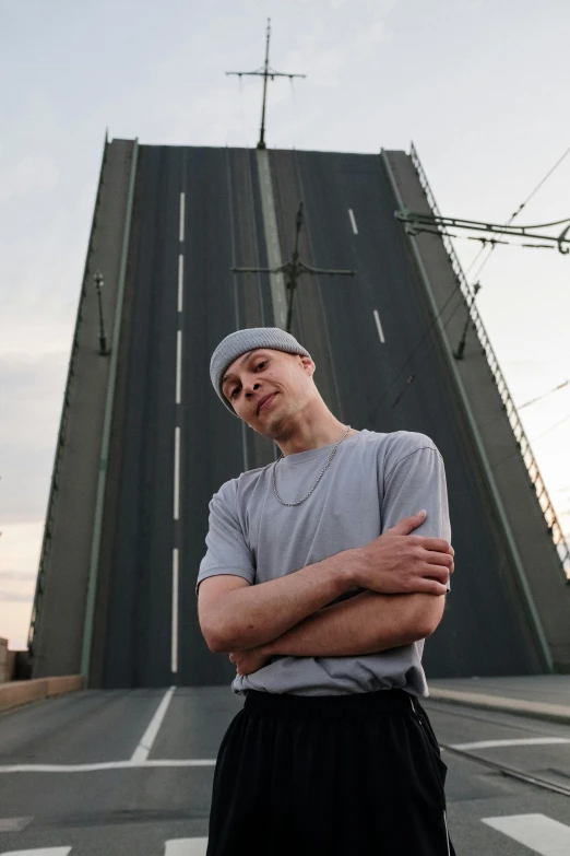 a man standing in front of a bridge, an album cover, inspired by Alexey Venetsianov, he also wears a grey beanie, standing on a skyscraper rooftop, 2019 trending photo, thin young male