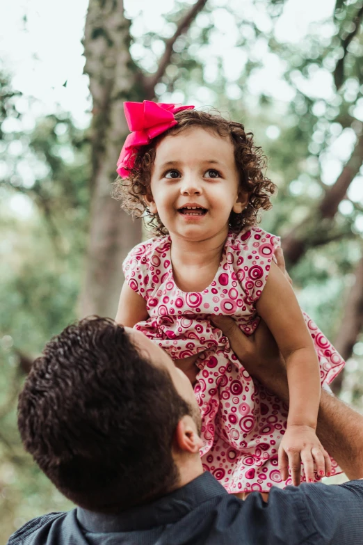 a man holding a little girl up in the air, a portrait, pexels contest winner, wearing a pink head band, sitting in a tree, hispanic, patterned clothing