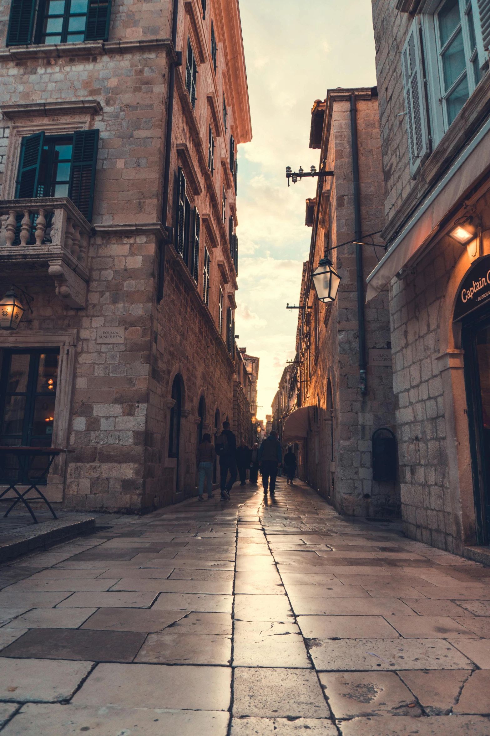 a couple of people walking down a street next to tall buildings, pexels contest winner, gothic quarter, sun down, buildings carved out of stone, pasta