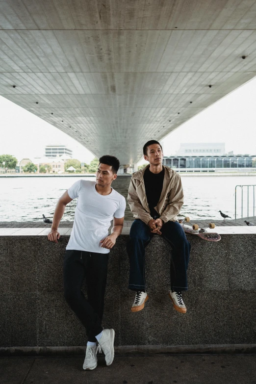 two men sitting on a ledge next to a body of water, by Niko Henrichon, asian man, concert, confident pose, bridges