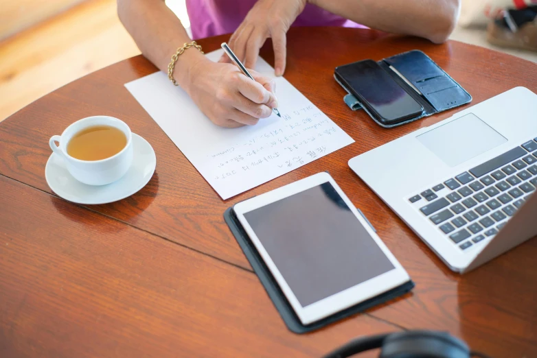 a woman sitting at a table writing on a piece of paper, trending on pexels, high-tech devices, table in front with a cup, raphael lecoste, avatar image