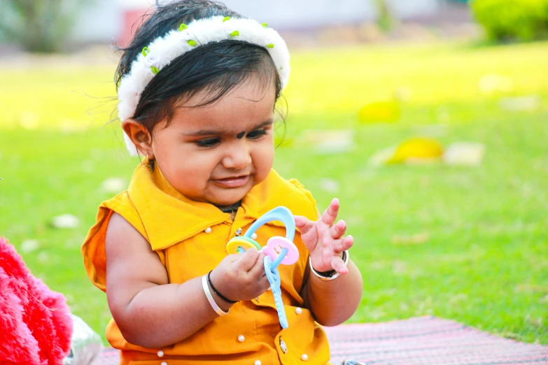 a baby sitting on a blanket playing with a toy, by Thota Vaikuntham, pexels contest winner, holding a bow, avatar image, at a park, wearing jewellery