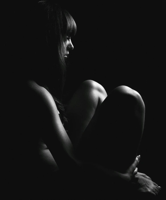 a woman sitting in the dark with her legs crossed, a black and white photo, unsplash, alternate album cover, abused, embracing, back light