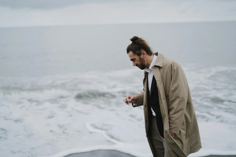 a man standing on top of a beach next to the ocean, light brown trenchcoat, windy hair, jovana rikalo, thumbnail