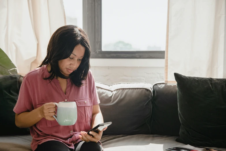 a woman sitting on a couch looking at her cell phone, pexels contest winner, with a white mug, asian female, dwell, next to a cup