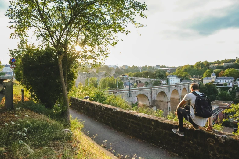 a man sitting on top of a stone wall, liege, aqueduct and arches, nature growing around the city, glowy light