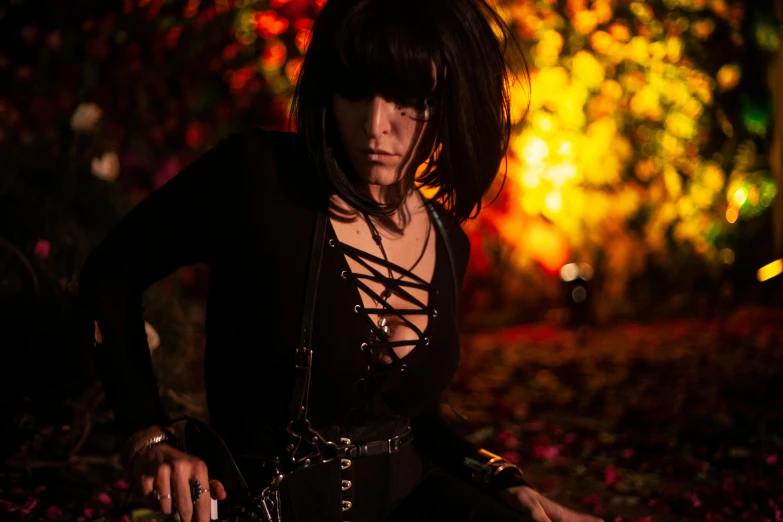 a woman sitting on the ground with a knife in her hand, an album cover, unsplash, gothic art, cosplayer, lights, black leather harness, amongst foliage