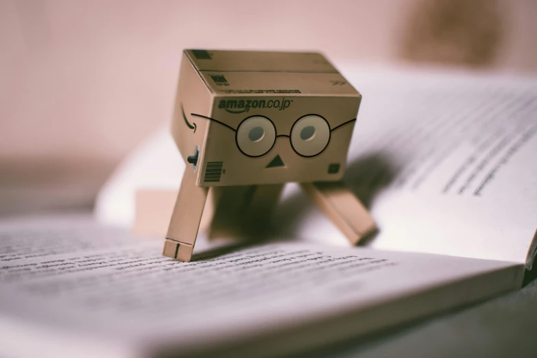 a cardboard robot sitting on top of an open book, pexels contest winner, square face, amateur photograph, performing, william gibson