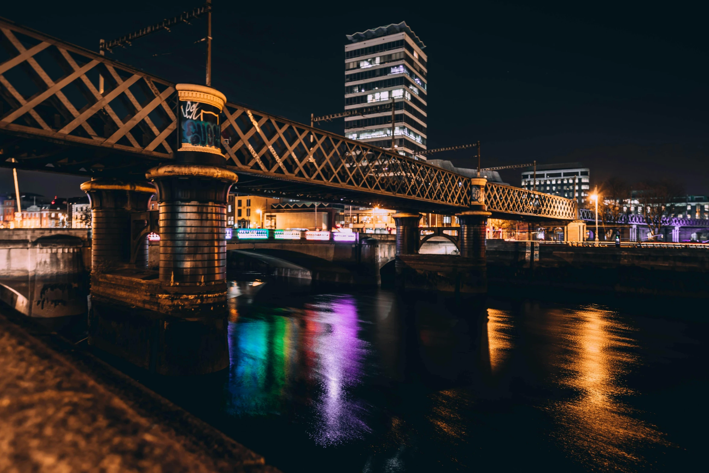 a bridge over a body of water at night, a picture, by Julia Pishtar, unsplash contest winner, graffiti, coventry city centre, colorful lights, manchester, multiple stories