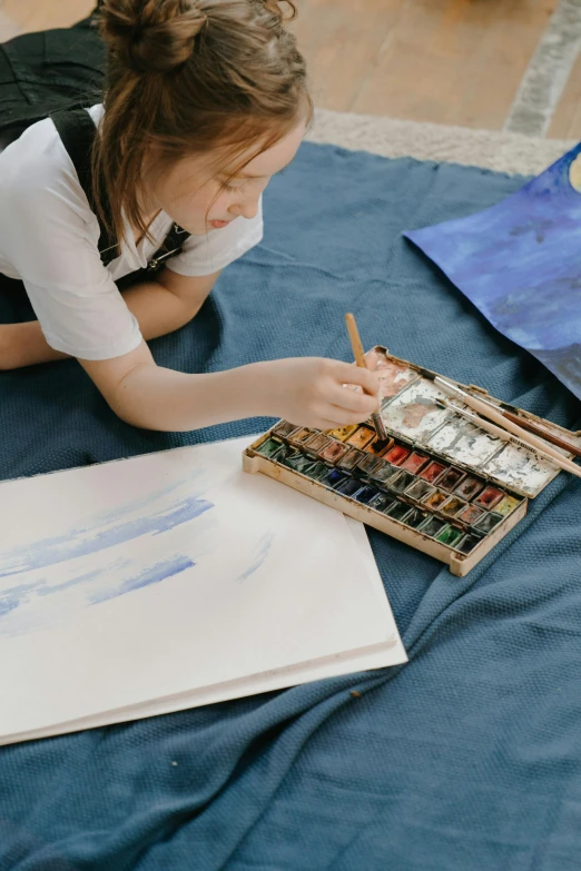 a little girl laying on the floor drawing on a piece of paper, inspired by artist, pexels contest winner, paintbrush and palettes, burnt umber and blue, painting on a canvas, painting on a badge