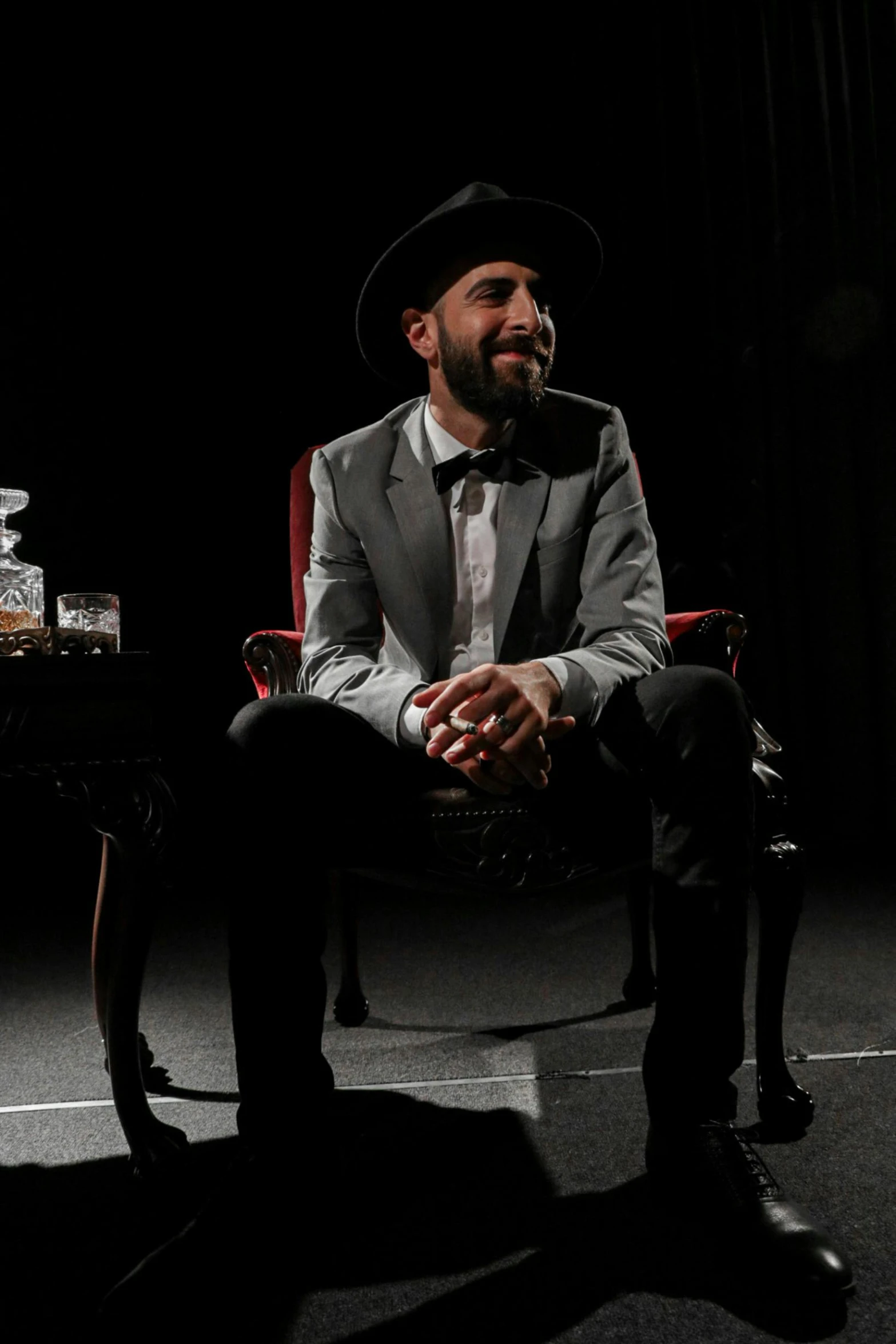 a man sitting in a chair in a dark room, a portrait, inspired by Germán Londoño, magician dog performing on stage, press shot, background : diego fazio, a suited man in a hat