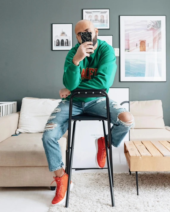 a man sitting on a chair taking a picture of himself, inspired by Jean-Yves Couliou, trending on pexels, maximalism, long orange sweatshirt, green legs, ripped jeans, bald man