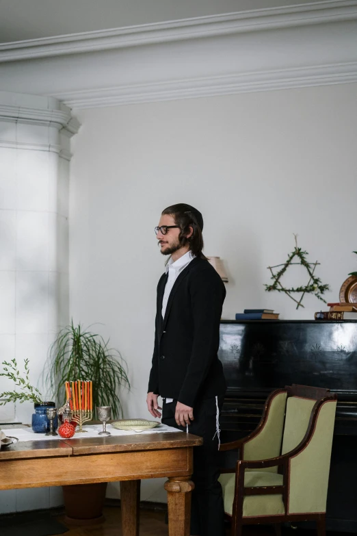 a man standing in a living room next to a piano, an album cover, unsplash, renaissance, jewish young man with glasses, at a dinner table, three piece suit, standing in front of the altar