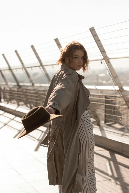 a woman standing on a bridge holding a hat, trending on pexels, renaissance, light brown trenchcoat, lorde, rooftop, musician