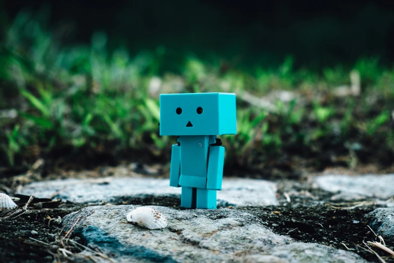 a small blue robot sitting on top of a rock, unsplash, pixel art, square face, toys, blurred photo, teal